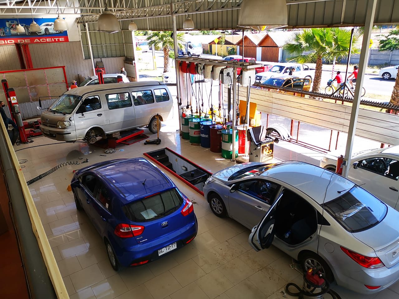 Interior del lubricentro: furgón en elevador, autos en las bahías y tambores de aceite con mangueras de dispensado.