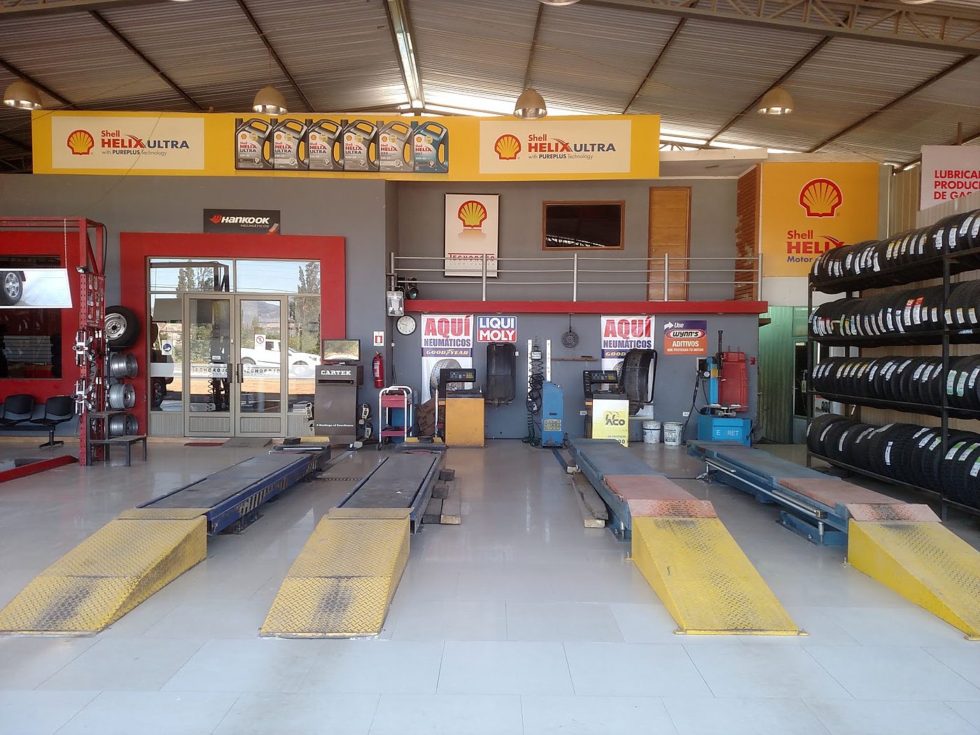 Góndola de neumáticos, rampas amarillas, máquina de alineación Cartek y carteles Aqui neumáticos, Hankook y Shell Helix Ultra.
