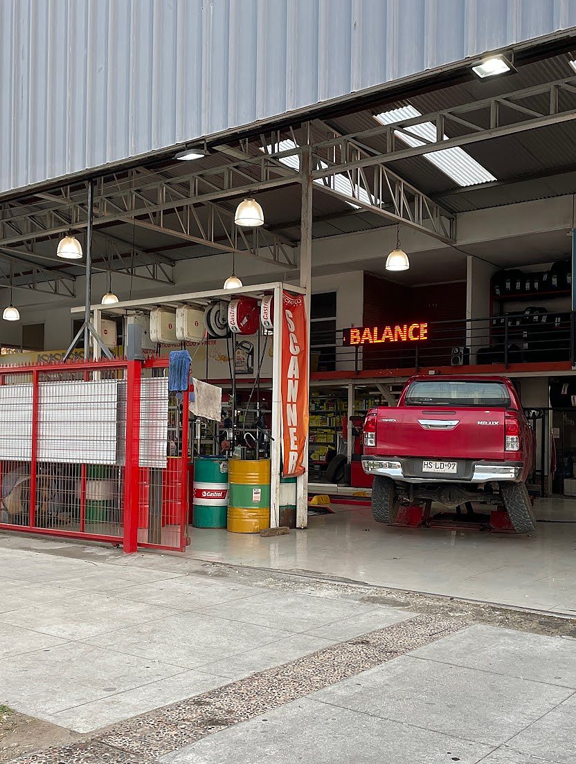 Entrada del taller con reja roja, tambores Castrol, pendón Scanner, letrero Balance y pickup en el elevador.